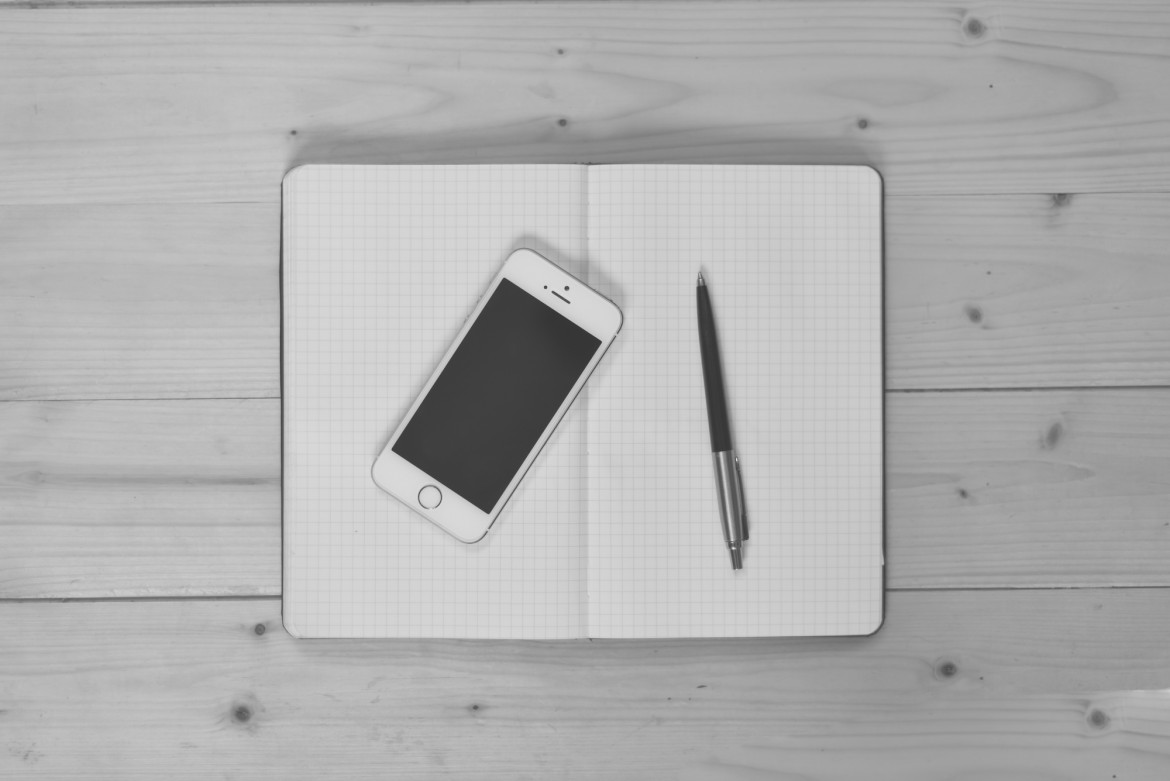 Overhead view of a smartphone and pen resting on an open blank notebook on a wooden table, black and white