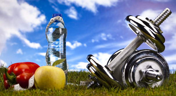 Fitness and health themed still life with dumbbells, a water bottle, measuring tape, and fresh produce on grass under natural sunlight